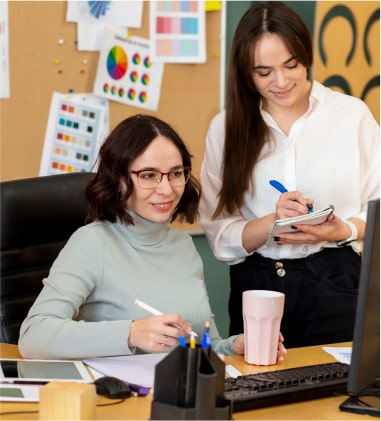 Two women collaborating on a project at a desk in a modern office setting.